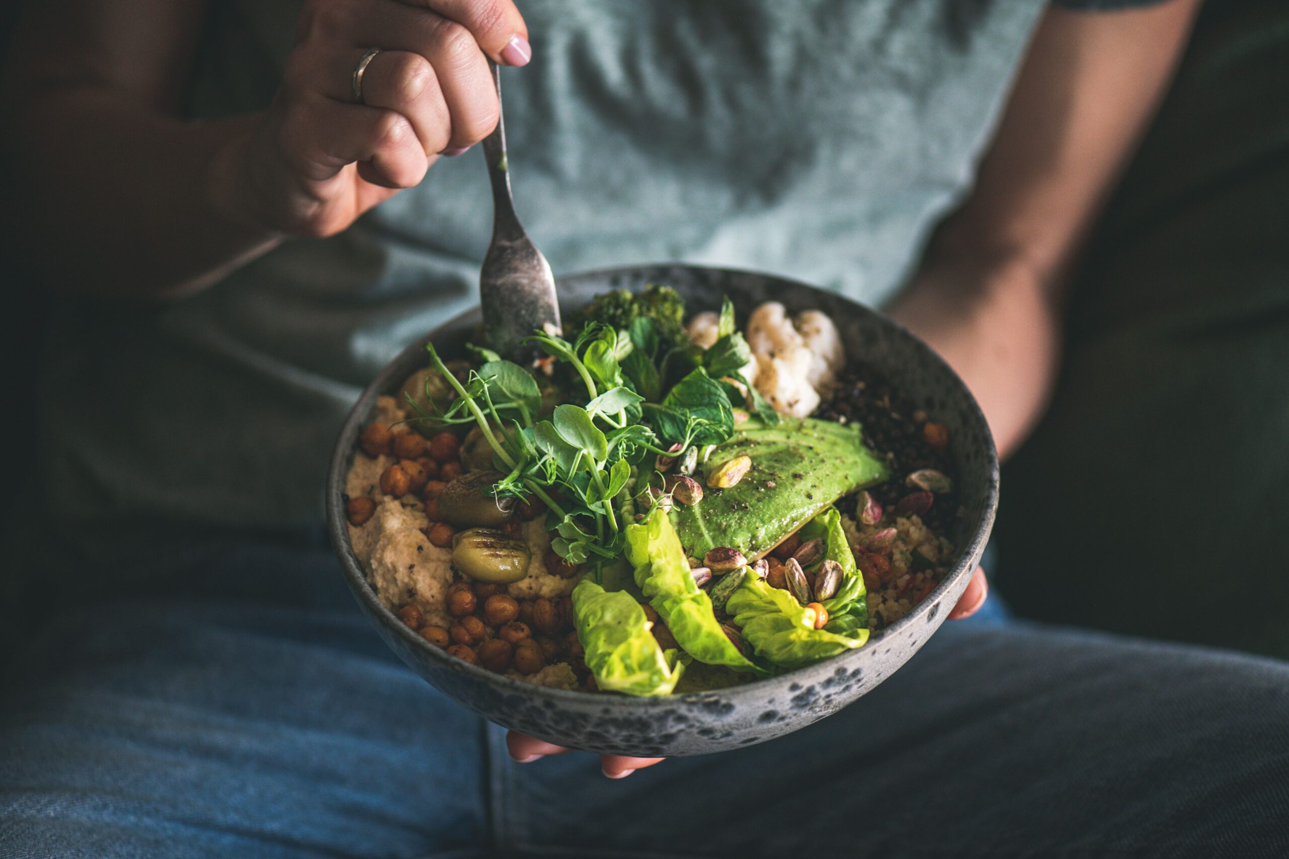 Gesundes Abendessen oder Mittagessen. Frau in T-Shirt und Jeans isst vegane Superbowl oder Buddha Bowl mit Hummus, Gemüse, Salat, Bohnen, Couscous und Avocado